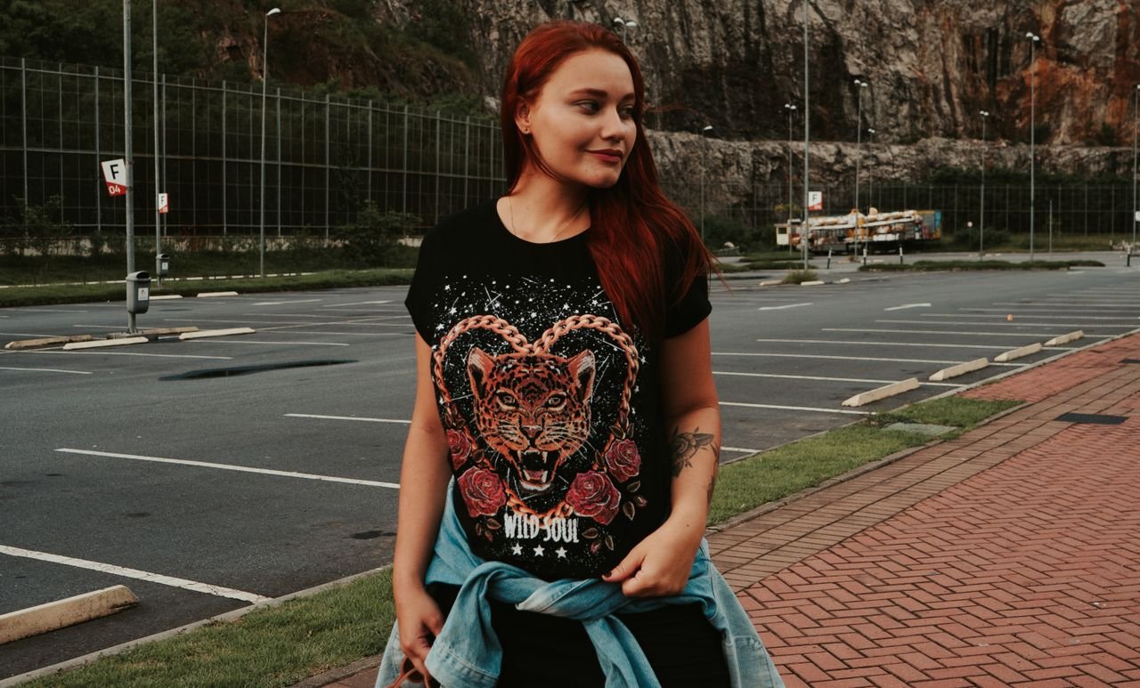 Portrait of a redhead young woman in a bold graphic t-shirt, standing in a parking lot with a relaxed expression.